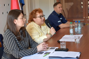 W centrum kadru, przy stole siedzą trzy osoby. Od prawej, w dalszym planie - umundurowany policjant, w środku kobieta w jasnej bluzce, trzyma w rękach mikrofon, po lewej w najbliższym planie, kobieta w czarno-białej bluzce.