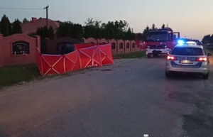 miejsce wypadku, na drodze widoczny radiowóz z włączoną sygnalizacją, oraz wóz strażacki, po lewej stronie murowane ogrodzenie, przed nim czerwony parawan z napisem straż