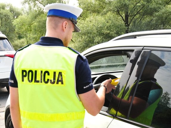 policjant ruchu drogowego w białej czapce oraz żółtej kamizelce z napisem policja wykonuje sprawdzenie stanu trzeźwości kierującego pojazdem