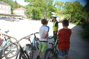 Zdjęcie kolorowe wykonane w porze dziennej przedstawia grupę dzieci która w obecności policjanta wydziału ruchu drogowego zdaje egzamin na kartę rowerową.