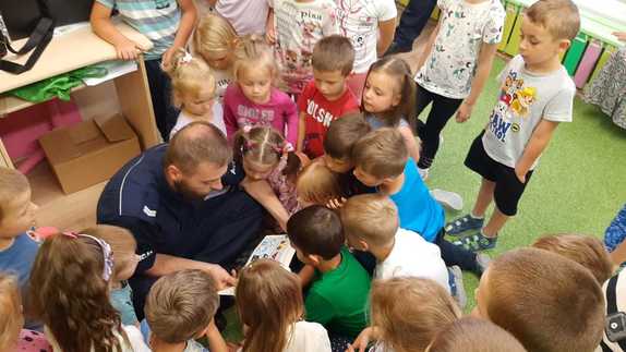 Zdjęcie kolorowe wykonane w porze dziennej przedstawia grupę przedszkolaków która siedzi na dywanie w sali i słucha prelekcji policjanta na temat bezpieczeństwa podczas wypoczynku nad wodą