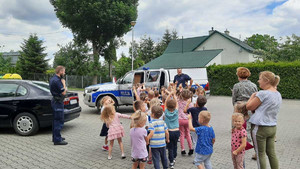 Zdjęcie kolorowe wykonane w porze dziennej przedstawia grupę przedszkolaków która stoi przed radiowozem  i słucha  policjanta który opowiada przedszkolakom o pracy jaka wykonuje na co dzień