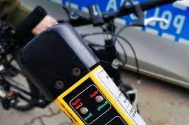Zdjęcie kolorowe przedstawia uradzenie do pomiaru alkoholu w wydychanym powietrzu tz. Alco-Blov w kolorze żółtym z czarnym ustnikiem. Urządzenie jest sfotografowane na tle kierownicy roweru a w oddali widoczna jest część radiowozu policyjnego oznakowanego