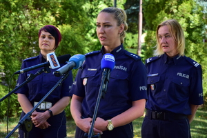 Trzy policjantki podczas briefingu. Stojąca w środku mówi do mikrofonów. W tle zieleń - drzewa i trawa