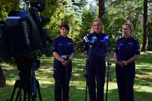 Trzy policjantki podczas briefingu. Stojąca w środku mówi do mikrofonów. W tle zieleń - drzewa i trawa