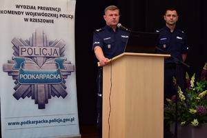 Policjant na mównicy przy mikrofonie. Po prawej drugi policjant. W tle banner z policyjną gwiazdą i napisem Wydział Prewencji Komendy Wojewódzkiej Policji w Rzeszowie, napis POLICJA PODKARPACKA oraz www.podkarpacka.policja.gov.pl.