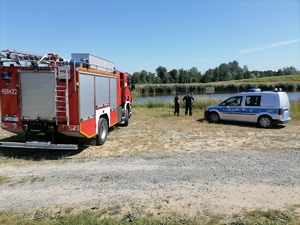Policjant wraz z strażakiem stoi przed zbiornikiem wodnym. Obok znajduje się oznakowany radiowóz i wóz strażacki.