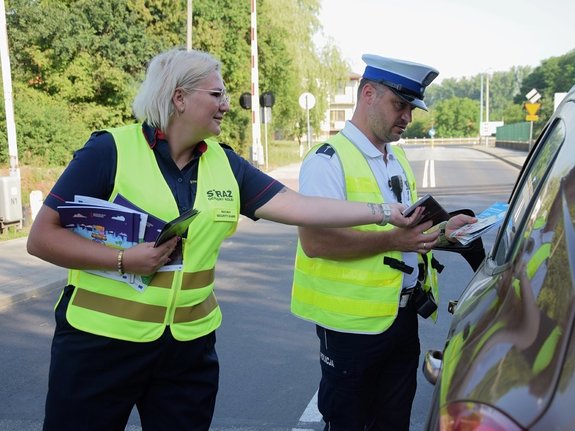 policjant ruchu drogowego w żółtej kamizelce oraz funkcjonariuszka straży ochrony kolei przekazują kierującemu pakiet ulotek informacyjnych