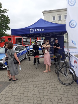 Na zdjęciu znajduje się rower policyjny, radiowóz, policjanci stojący pod namiotem oraz kobiety odwiedzające policyjne stoisko