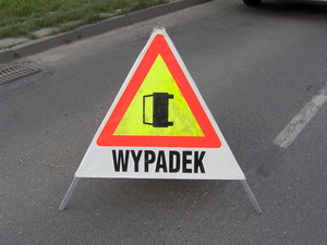 Na zdjęciu znajduje się trójkątny znak drogowy ustawiony na jezdni z napisem wypadek