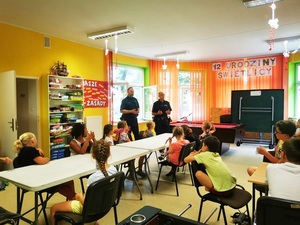 policjant i strażnik miejski podczas prelekcji dla dzieci. Na pierwszym planie siedzące na krzesełkach dzieci słuchające wykładu