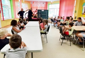 policjant i strażnik miejski podczas prelekcji dla dzieci, wskazując na elementy wyposażenia roweru. Na pierwszym planie siedzące na krzesełkach dzieci słuchające wykładu