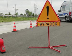 droga, na niej znak ostrzegawczy z napisem wypadek, w tle radiowóz