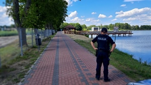 Na zdjęciu znajduje się policjant stojący na chodniku nad brzegiem zalewu wodnego