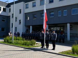 na zdjęciu policjanci z pocztu flagowego stojący przy maszcie przygotowani do podniesienia flagi państwowej, w tle budynek komendy policji oraz stojący w szyku policjanci