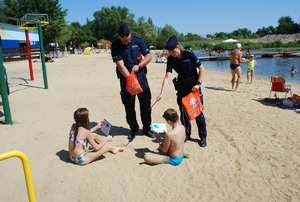 Kąpielisko &quot;Żwirownia&quot;. Na plaży ubrani w niebieskie mundury policjantka i policjant wręczają siedzącej na piasku dwójce dzieci broszurki profilaktyczne i elementy odblaskowe.