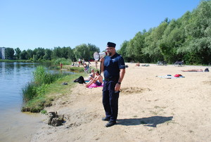 Kąpielisko &quot;Żwirownia&quot;, na zdjeciu policjant w granatowym mundurze, stojący na brzegu kąpieliska poprzez megafon prosi kapiących się o korzystanie z miejsca wyznaczonego do kąpieli. W tle widać plaże i wypoczywające osoby.