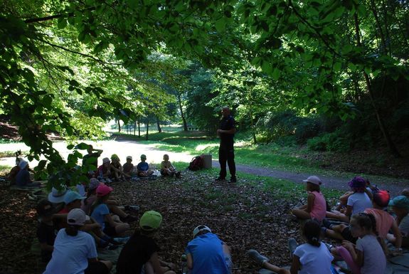Policjant z dziećmi podczas prelekcji. Spotkanie odbywa się w parku.
