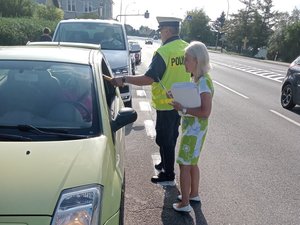 umundurowany policjant stojący przy samochodzie wykonuje sprawdzenie stanu trzeźwości kierującego, obok kobieta (członek MKRPA) trzymająca broszury i ulotki