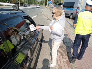 kobieta (członek MKRPA) przekazująca kierującemu broszury i ulotki w tle odchodzący od samochodu policjant w żółtej kamizelce