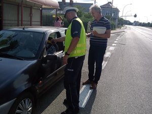 umundurowany policjant stojący przy samochodzie wykonuje sprawdzenie stanu trzeźwości kierującego, obok członek MKRPA w Jaśle z pakietem ulotek