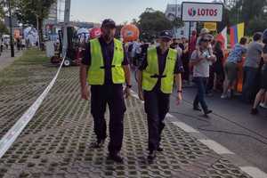 Dwoje policjantów, w tle kibice stojący wzdłuż ogrodzenia trasy wyścigu kolarskiego.