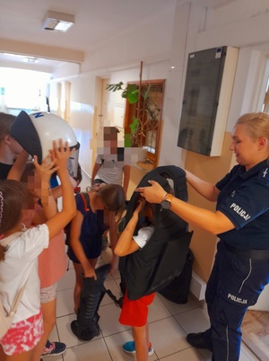 na zdjęciu policjantka z policjantem stoja z dziećmi na korytarzu. jedno z dzieci ubiera kamizelkę przeciwuderzeniową