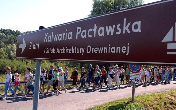 Zdjęcie kolorowe przedstawia znak drogowy w kolorze granatowym na którym widnieje napis w białym kolorze „Kalwaria Pacławska 2 km –szlak architektury drewnianej” po lewej stronie znaku widoczni są pielgrzymi.