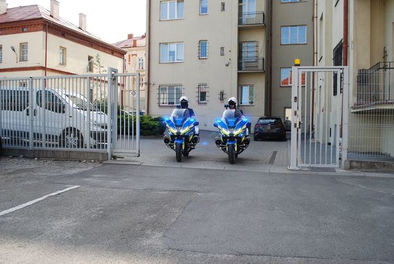 Na zdjęciu dwaj policyjni motocykliści wyjeżdżają na patrol z dziedzińca KMP. Widać nowe motory bmw w nowych barwach w koralach żółtym, srebrnym i niebieskim. W tel dziedziniecc komendy widziany przez uchylona bramę.