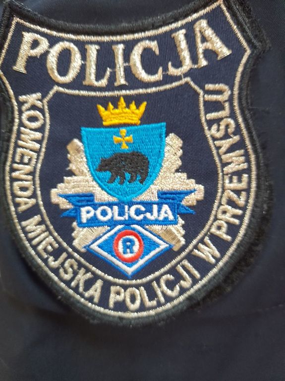 Zdjęcie przedstawia emblemat policjanta ruchu drogowego. Na górze napis POLICJA w środku logo miasta Przemyśla (niedźwiedź koloru czarnego a nad nim krzyż oraz korona w kolorze złotym , na obrzeżach emblematu widnieje napis Komenda Miejska Policji w Przemyślu