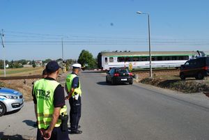 W rejonie przejazdu kolejowego widoczny pojazd, który zatrzymał się w związku z przejazdem pociągu. Na poboczu stoi funkcjonariusz policji oraz dwóch funkcjoanriusze służby ochrony kolei.