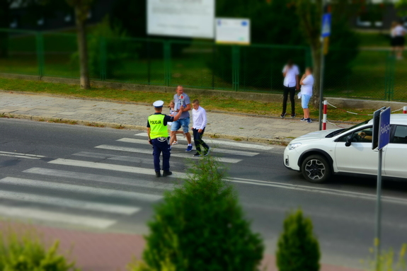 Policjant stojący na przejściu dla pieszych, trzymający tarczę do zatrzymywania pojazdów,ułatwia w ten sposób przejście dzieci przez jezdnię.
