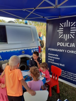 Na zdjęciu znajduje się radiowóz, policyjny baner i policjantka z dziećmi