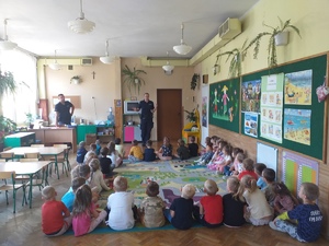 Zdjęcie kolorowe przedstawia sala przedszkolna w której na dywanie siedzą dzieci a przed nimi stoi umundurowana policjantka wraz z policjantem.