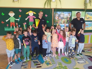 Zdjęcie kolorowe przedstawia salę przedszkolna w której dzieci przedszkolne wraz z policjantem i policjantka stoją i pozują do zdjecia