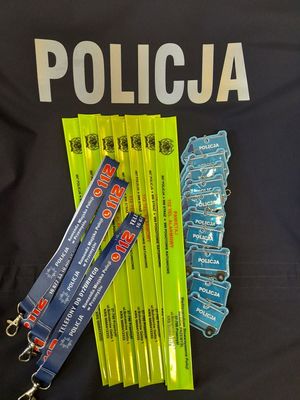 Zdjęcie kolorowe przedstawia elementy odblaskowe (opaski) w kolorze żółtym oraz breloczki niebieskie w kształcie radiowozu policyjnego . Odblaski leżą na ciemnym tle na którym widnieje napis POLICJA . Po lewej stronie zdjęcie na odblaskach leży trzy smyczki z logo Komendy Miejskiej Policji w Przemyślu