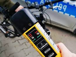 Zdjęcie kolorowe przedstawia urządzenie do pomiaru alkoholu w wydychanym powietrzu „Alcoblov” w żółtym kolorze z czarnym ustnikiem które jest trzymane w reku. W tel widoczny jest rower oraz część radiowozu policyjnego oznakowanego