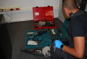 Na zdjęciu policjant w  cywilnym ubraniu, w czarnej kamizelce, kuca przy  leżących na podłodze trzech walizkach z elektrosprzętem.