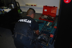 Na zdjęciu odwrócony policjant w ubiorze cywilnym, w czarnej kamizelce z napisem policja na plecach. Przed policjantem na podłodze leżą w walizkach elektronarzędzia.