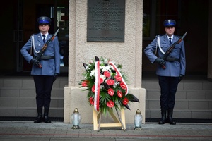 Policyjna warta honorowa przy tablicy pamiątkowej umieszczonej na kolumnie przed wejściem do komendy. Pod tablicą na drewnianym stojaku złożony wieniec. Na ziemi stoją dwa znicze.