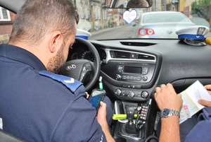 Zdjęcie kolorowe wykonane w porze dziennej przedstawia wnętrze radiowozu policyjnego w którym siedzi dwóch f-szy policji i wykonują czynności sprawdzające w stosunku do kierującego pojazdem vw którego zatrzymano do kontroli drogowej