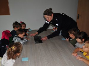 na zdjęciu siedzące za stołem dzieci odbijają swoje odciski palców na karteczkach papieru, pomaga im umundurowana policjantka