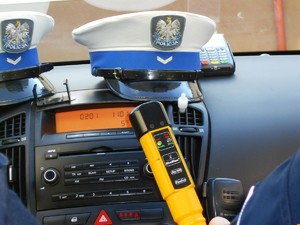 Czapka policjanta ruchu drogowego leżąca na podszybiu radiowozu, przed nią trzymane przez policjanta urządzenie do badania stanu trzeźwości.