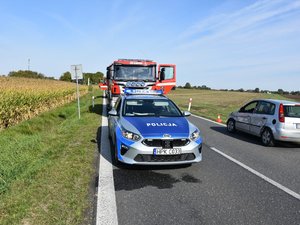 Na drodze wojewódzkiej stoi oznakowany radiowóz. Za nim znajduje się wóz strażacki obok natomiast pojazd marki ford z widocznym uszkodzeniem całej lewej strony pojazdu.