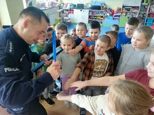 Zdjęcie kolorowe wykonane w porze dziennej –przedstawia grupę dzieci wraz z policjantem w sali szkolnej