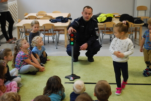 Policjant omawia z dziećmi zasady bezpieczeństwa ruchu drogowego. W tym celu wykorzystuje mini sygnalizator świetlny.
