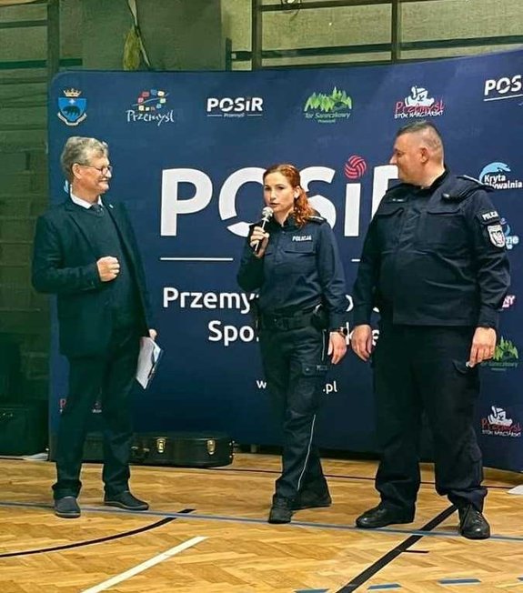 Zdjęcie kolorowe przedstawia halę Przemyskiego Ośrodka Sportu i Rekreacji w której stoi dwóch funkcjonariuszy policji wspólnie z konferansjerem. Policjantka trzyma w ręku mikrofon prowadzi wykład dla seniorów
