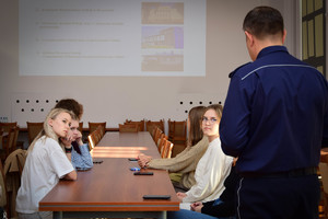 Policjant podczas spotkania ze studentami w auli komendy. Policjant stoi, studenci siedzą na krzesłach przy stole.