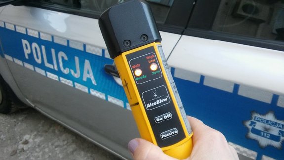 Zdjęcie przedstawia urządzenie do pomiaru alkoholu w wydychanym powietrzu „Alcoblov” w kolorze żółtym z czarnym ustnikiem , które jest trzymane w ręku. W tle widoczny jest lewy bok pojazdu policyjnego oznakowanego na którym widnieje napis POLICJA w białym kolorze na niebieskim tle