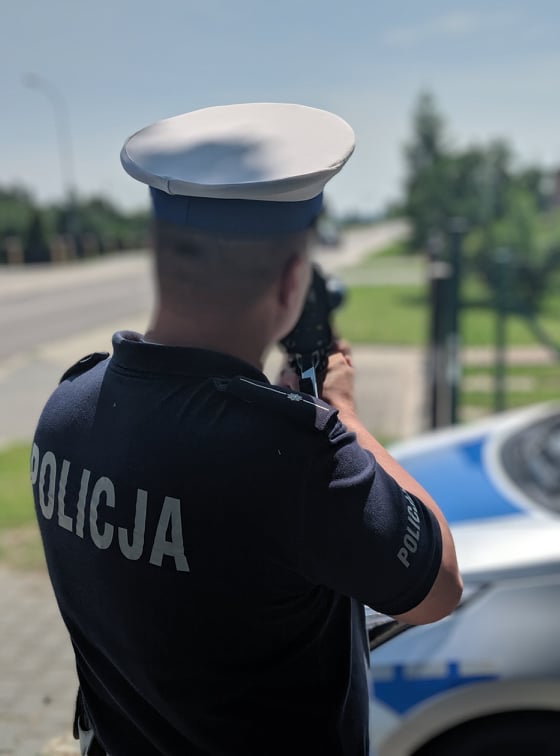 Policjant ruchu drogowego w białej czapce mierzący prędkość jadącego samochodu, w tle radiowóz.